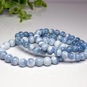 May include: Two stretch bracelets composed of smooth, round beads in varying shades of light blue, medium blue, and white. The beads have a polished, semi-translucent finish with swirling patterns, arranged in a continuous circular loop design.