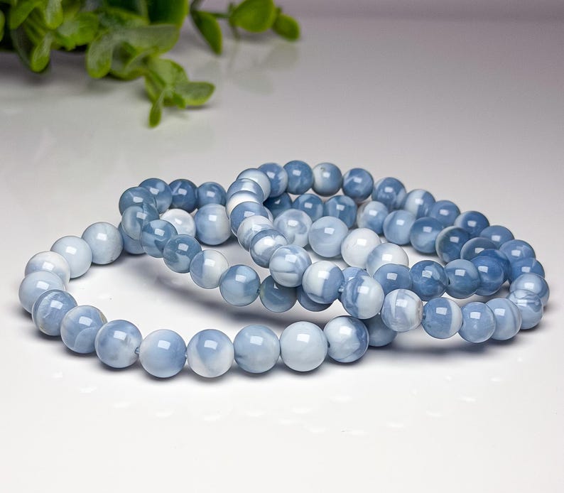 May include: Two stretch bracelets composed of smooth, round beads in varying shades of light blue, medium blue, and white. The beads have a polished, semi-translucent finish with swirling patterns, arranged in a continuous circular loop design.