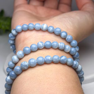 May include: A multi-strand bracelet composed of smooth, round, light blue beads with subtle white marbled patterns. The beads are strung together in a continuous loop, creating a layered appearance when wrapped around the wrist multiple times.