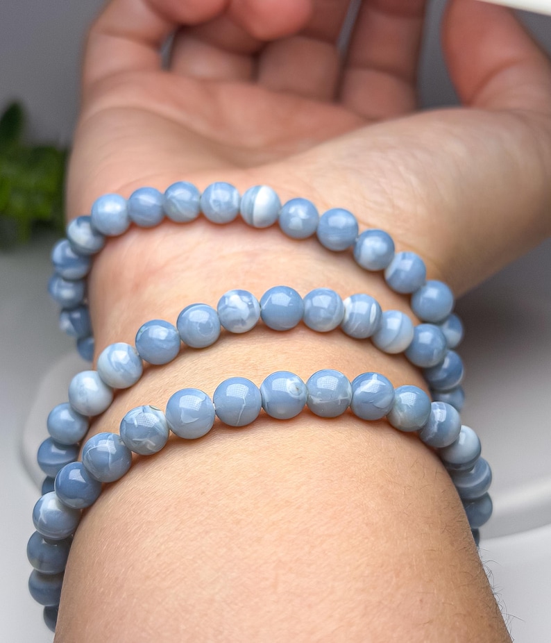 May include: A multi-strand bracelet composed of smooth, round, light blue beads with subtle white marbled patterns. The beads are strung together in a continuous loop, creating a layered appearance when wrapped around the wrist multiple times.