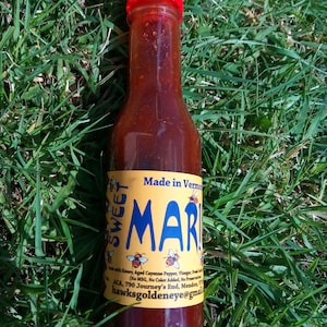 May include: A glass bottle of Sweet Mari hot sauce with a red cap. The label is yellow with black text that reads "Sweet Mari" and "Made in Vermont". The label also includes ingredients and contact information.