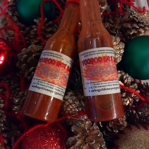 May include: Two small glass bottles of red hot sauce with a Christmas-themed background. The bottles are labeled "Voodoo Hot Sauce" and contain a blend of aged cayenne peppers, vinegar, and other ingredients. The bottles are surrounded by pine cones, red and green ornaments, and red string.
