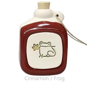 May include: A ceramic jug with a red-brown exterior and a white center panel featuring a frog holding a flower. The jug has a cork stopper and a string for hanging. The text "Cinnamon / Frog" is at the bottom.