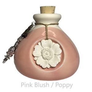 May include: A rounded ceramic bottle with a glossy pink blush finish, featuring a white sculpted poppy flower relief on the front. The bottle has a cork stopper, a twine tie, and a decorative silver-toned metal charm with beads hanging from the neck.