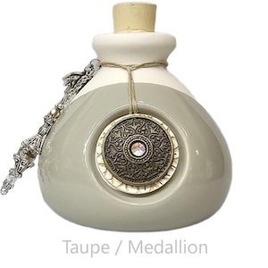 May include: A taupe-colored ceramic bottle with a white top and a cork stopper. A decorative medallion with a central crystal and intricate design hangs from a twine. A silver chain with decorative elements is attached to the bottle.