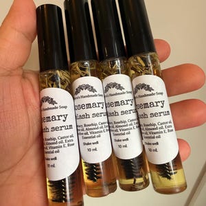 May include: Four small glass bottles of rosemary lash serum are held in a hand. Each bottle has a black cap and a white label with text. The serum is amber-colored, and a brush applicator is visible inside.