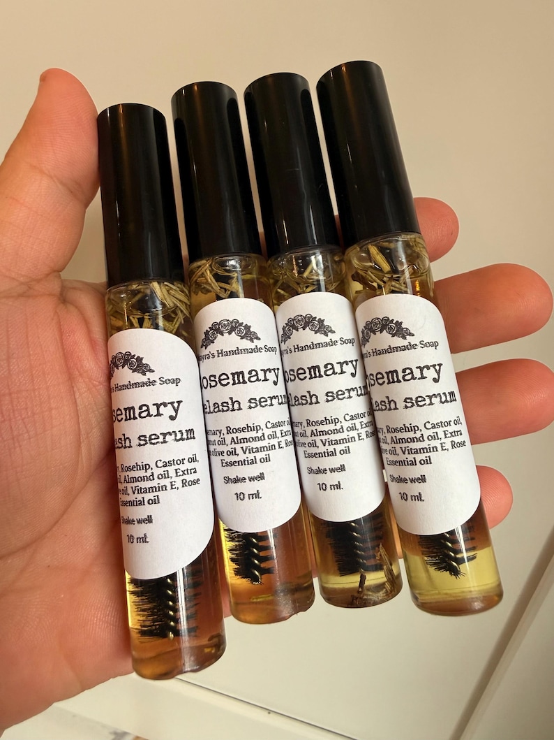 May include: Four small glass bottles of rosemary lash serum are held in a hand. Each bottle has a black cap and a white label with text. The serum is amber-colored, and a brush applicator is visible inside.