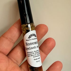 May include: A clear glass bottle of rosemary eyelash serum with a black cap. The label reads "Rosemary Eyelash Serum" and lists ingredients like rosemary, coconut oil, and almond oil. The bottle contains 10 ml of product.