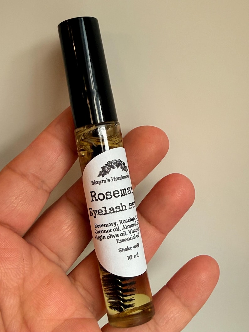 May include: A clear glass bottle of rosemary eyelash serum with a black cap. The label reads "Rosemary Eyelash Serum" and lists ingredients like rosemary, coconut oil, and almond oil. The bottle contains 10 ml of product.