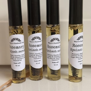 May include: Four clear glass roller bottles with black caps, filled with a yellow liquid and rosemary sprigs. Each bottle has a white label with black text that reads "Rosemary Eyelash Serum" and "10 ml".