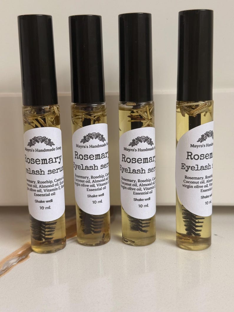 May include: Four clear glass roller bottles with black caps, filled with a yellow liquid and rosemary sprigs. Each bottle has a white label with black text that reads "Rosemary Eyelash Serum" and "10 ml".