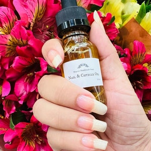 May include: A small glass bottle of "Nail & Cuticle Oil" with a black dropper top is held in front of a bouquet of pink and yellow flowers. The bottle has a white label with the product name and brand. The person's nails are long and painted.