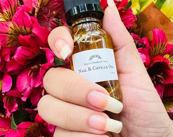 May include: A small glass bottle of "Nail & Cuticle Oil" with a black dropper top is held in front of a bouquet of pink and yellow flowers. The bottle has a white label with the product name and brand. The person's nails are long and painted.
