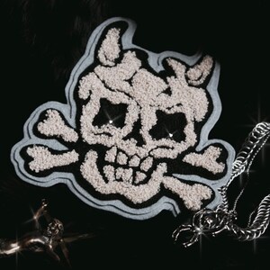 Op de afbeelding: Een wit geborduurde schedel en gekruiste beenderen patch met een blauwe achtergrond. De patch ligt op een zwart oppervlak.