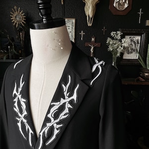 May include: A black blazer with white embroidered designs on the collar and shoulders. The blazer is on a mannequin in a room with a dark background.