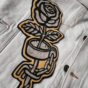 K&ouml;nnte beinhalten: Ein gestickter Patch in Schwarz und Grau mit einer Rose, um deren Stiel eine Kette gewickelt ist. Die Rose befindet sich auf einem braunen Hintergrund.