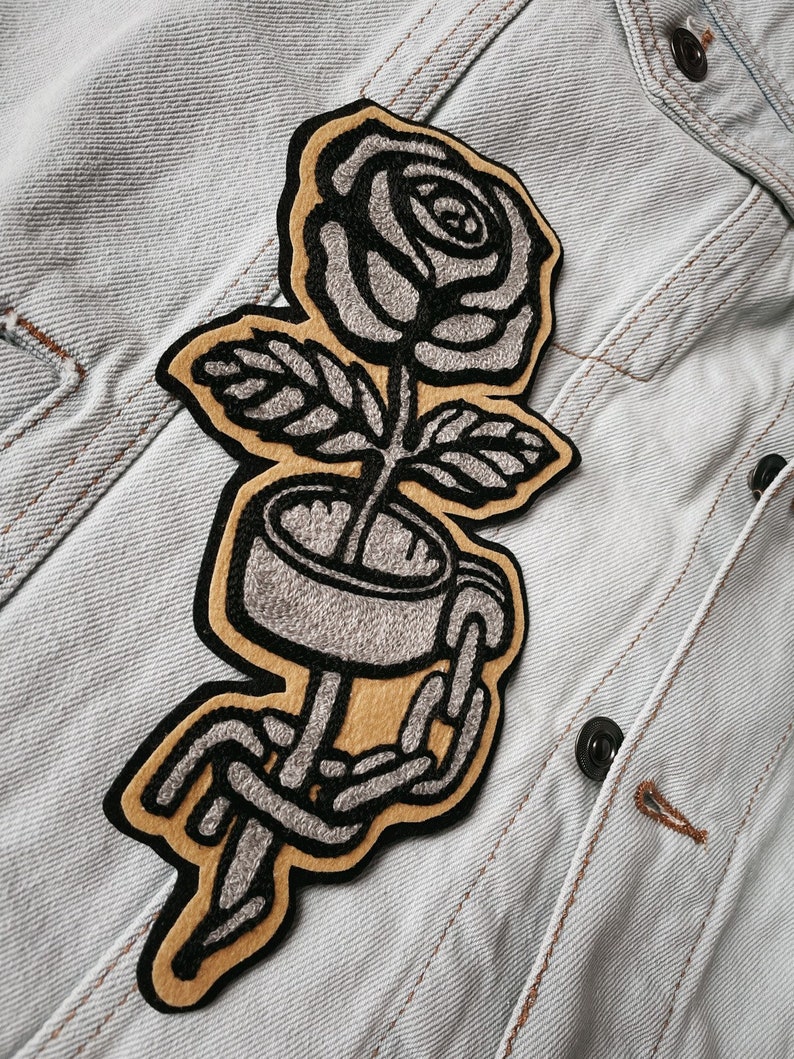 K&ouml;nnte beinhalten: Ein gestickter Patch in Schwarz und Grau mit einer Rose, um deren Stiel eine Kette gewickelt ist. Die Rose befindet sich auf einem braunen Hintergrund.