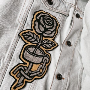 K&ouml;nnte beinhalten: Ein gestickter Aufn&auml;her in Schwarz und Grau, der eine Rose mit einer Kette um den Stiel zeigt. Der Aufn&auml;her ist auf eine hellblaue Jeansjacke gen&auml;ht.