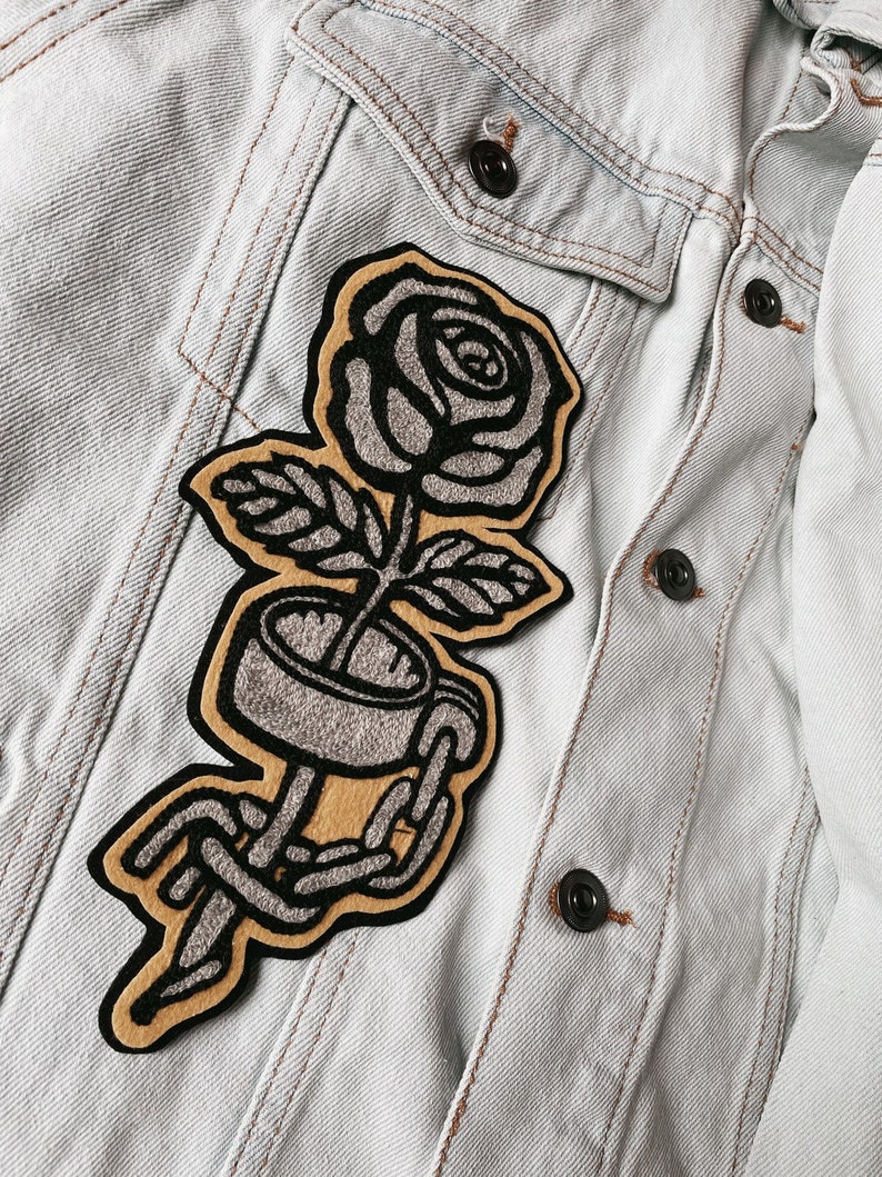 K&ouml;nnte beinhalten: Ein gestickter Aufn&auml;her in Schwarz und Grau, der eine Rose mit einer Kette um den Stiel zeigt. Der Aufn&auml;her ist auf eine hellblaue Jeansjacke gen&auml;ht.