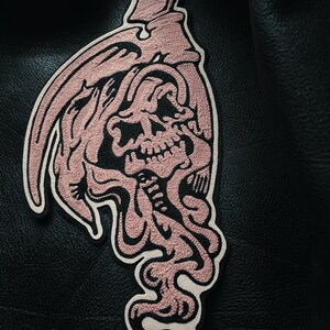 May include: A pink embroidered patch of a skull with a scythe, on a black leather jacket.