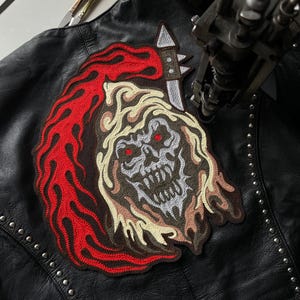 Pode incluir: Um remendo bordado em vermelho, branco e cinza de uma caveira com chamas e uma faca. O remendo está sendo costurado em uma jaqueta de couro preta.