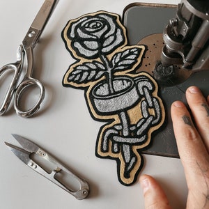 May include: A white embroidered rose with a chain wrapped around the stem, on a beige fabric patch. The patch is being sewn onto a sewing machine.