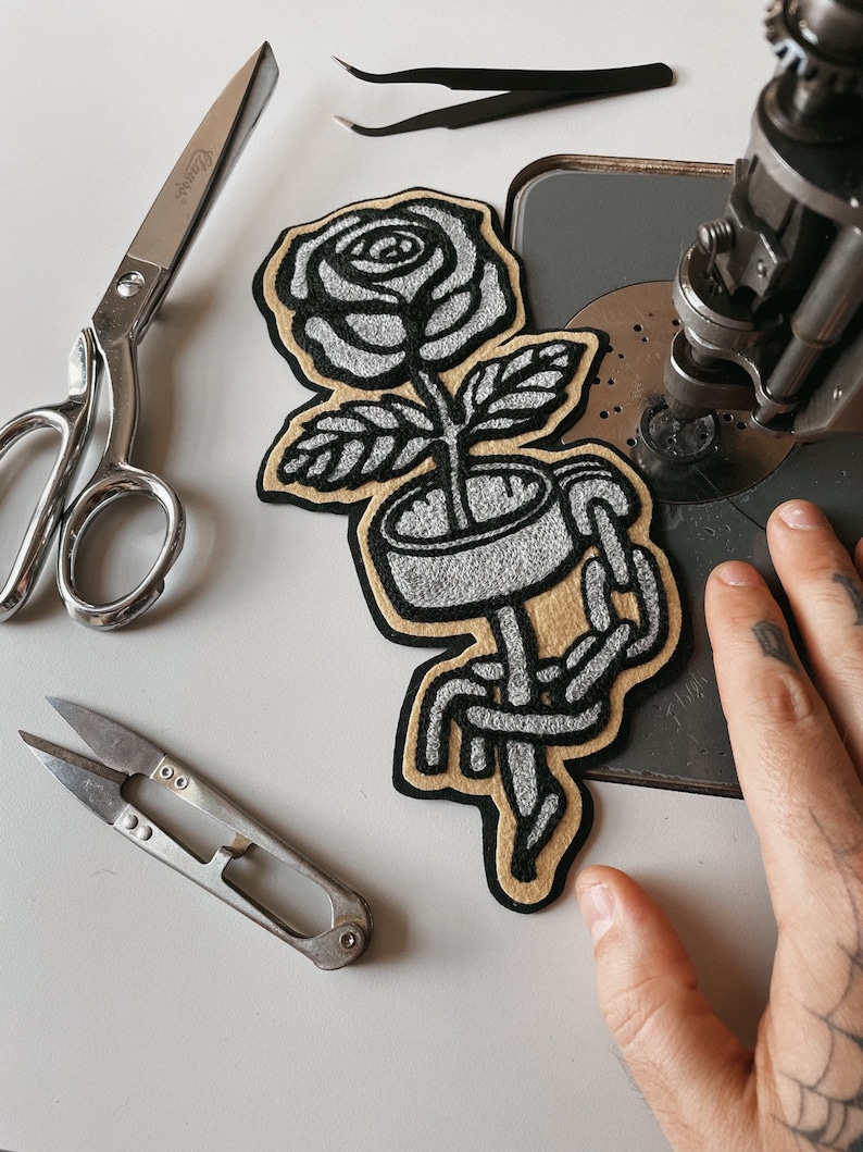 K&ouml;nnte beinhalten: Ein beigefarbener Stoff-Patch mit einer gestickten wei&szlig;en Rose, um deren Stiel eine Kette gewickelt ist. Der Patch wird auf einer N&auml;hmaschine befestigt.