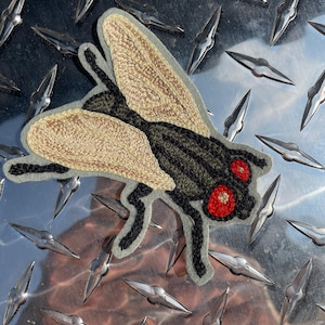 May include: A gray, black, and beige embroidered patch of a fly with red eyes. The fly is on a silver background.