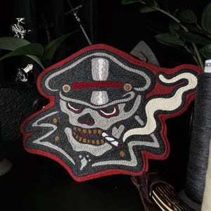 Puede incluir: Un parche bordado que representa una calavera con un sombrero militar fumando un cigarrillo. La calavera es roja, blanca y gris con un diente de oro y un bigote blanco.