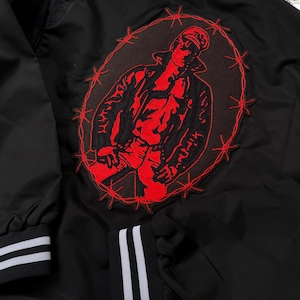 May include: Black bomber jacket with a red embroidered patch on the back. The patch features a figure in a leather jacket and a cap, surrounded by barbed wire.