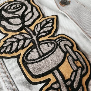 K&ouml;nnte beinhalten: Ein gestickter Patch in Schwarzwei&szlig;, der eine Rose in einer Tasse zeigt, die von skelettartigen H&auml;nden gehalten wird. Die Rose ist grau und die Tasse ist grau mit schwarzem Umriss. Die H&auml;nde sind grau mit schwarzem Umriss und halten die Tasse auf einem gelben Hintergrund.