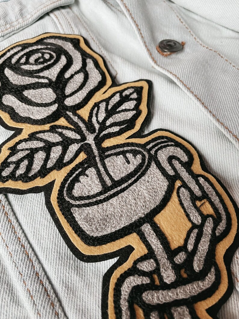 K&ouml;nnte beinhalten: Ein gestickter Patch in Schwarzwei&szlig;, der eine Rose in einer Tasse zeigt, die von skelettartigen H&auml;nden gehalten wird. Die Rose ist grau und die Tasse ist grau mit schwarzem Umriss. Die H&auml;nde sind grau mit schwarzem Umriss und halten die Tasse auf einem gelben Hintergrund.