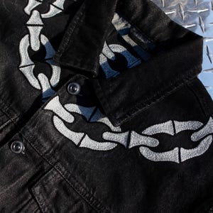 May include: Black denim jacket with a white chain bone design on the front. The jacket has a button closure and two pockets.