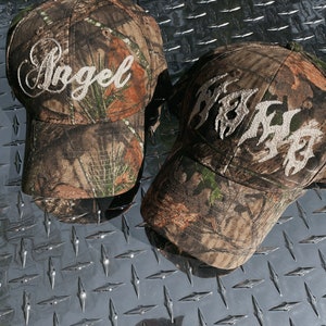 Peut inclure: Deux casquettes de baseball en camouflage avec du texte brodé blanc. La première casquette dit "Angel" et la deuxième casquette dit "Fresh".