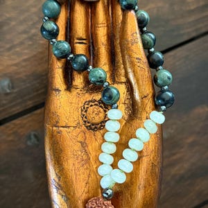 Può includere: Una collana mala con perline di pietra verde e nera, una pietra di colore chiaro e un nappa. La collana è esposta su una scultura di mano in legno con finitura dorata. La mala è un accessorio spirituale.
