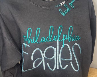 May include: Black sweatshirt with teal green embroidery that reads "Philadelphia Eagles" and "bird gang" on the front.