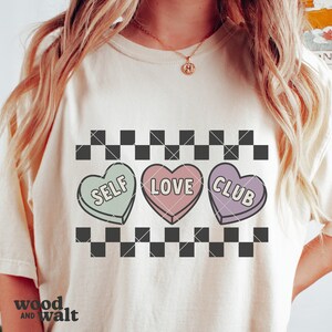 May include: A white t-shirt with a black and white checkered pattern and three candy hearts with the words "Self Love Club" on them.
