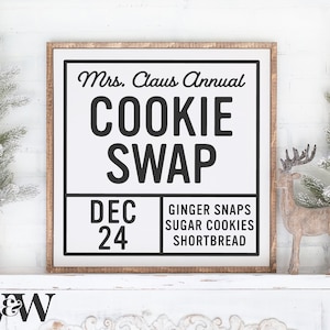 Puede incluir: Un letrero de madera blanco con texto negro que dice "Mrs. Claus Annual Cookie Swap Dec 24 Ginger Snaps Sugar Cookies Shortbread". El letrero está enmarcado en un marco de madera marrón claro y está sentado en un estante blanco con un fondo de ladrillo blanco.