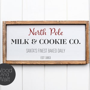 May include: A wooden sign with a white background and a brown frame. The sign reads "North Pole Milk & Cookie Co. Santa's Finest Baked Daily Est 1863".