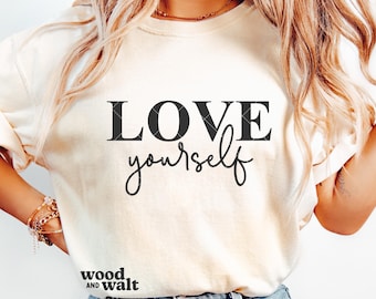 May include: A cream-colored t-shirt featuring the black text LOVE yourself printed on the front. The word LOVE is in a bold serif font, while yourself is in a cursive script. The text wood AND walt is printed in the bottom left corner.