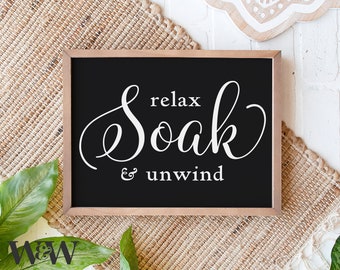 Może przedstawiać: Oprawiony czarny znak z białym napisem "relax Soak & unwind". Znak ma jasnobrązową drewnianą ramę i jest wyświetlany na tkanej macie. Słowa "relax" i "unwind" są w prostej czcionce, podczas gdy "Soak" jest w ozdobnym skrypcie.