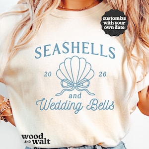 May include: A cream-coloured sweatshirt featuring the words "Seashells and Wedding Bells" in light blue, with a seashell graphic. The numbers "20" and "26" are on either side of the seashell. The words "wood and walt" are at the bottom.