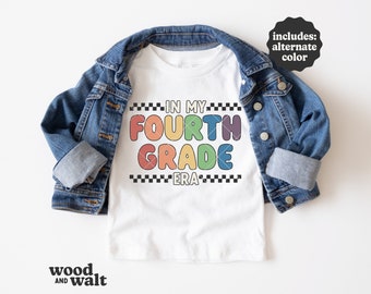 May include: A white t-shirt featuring the text IN MY FOURTH GRADE ERA in colorful bubble letters, bordered by black and white checkered lines, layered under a blue denim jacket. A black circular badge reads includes: alternate color.