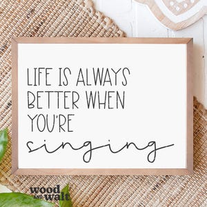 May include: A framed wooden sign with the text "LIFE IS ALWAYS BETTER WHEN YOU'RE singing" in black lettering. The sign has a white background and is set against a woven background.