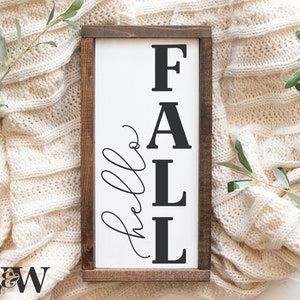 Puede incluir: Un letrero de madera blanco con la palabra "FALL" en letras negras y las palabras "hello fall" en escritura cursiva. El letrero está sobre una manta de punto blanca.