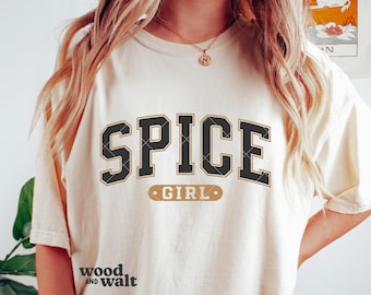 Puede incluir: Una camiseta de manga corta color blanco hueso con el texto SPICE en letras grandes negras de estilo universitario con un borde dorado fino, y la palabra GIRL en una insignia rectangular dorada más pequeña debajo. Tiene cuello redondo.