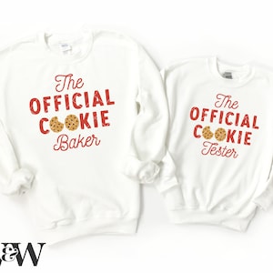 May include: Two white sweatshirts with red text and a cookie graphic. One sweatshirt says "The Official Cookie Baker" and the other says "The Official Cookie Tester".