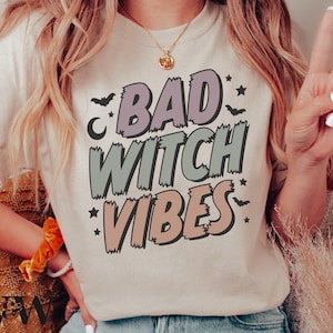 May include: A white t-shirt with a graphic print that says "Bad Witch Vibes" in a colorful, cartoon-style font. The graphic includes stars, crescent moons, and bats.
