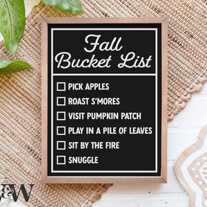 May include: A black chalkboard with a white wooden frame lists fall bucket list items. The list includes: pick apples, roast s'mores, visit pumpkin patch, play in a pile of leaves, sit by the fire, and snuggle.