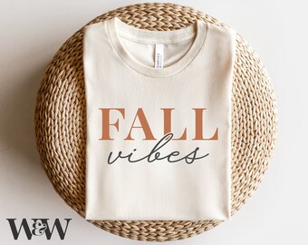 Puede incluir: Una camiseta blanca con el texto "FALL vibes" impreso en letras marrones y grises. La camiseta está doblada y colocada sobre una estera de paja tejida.